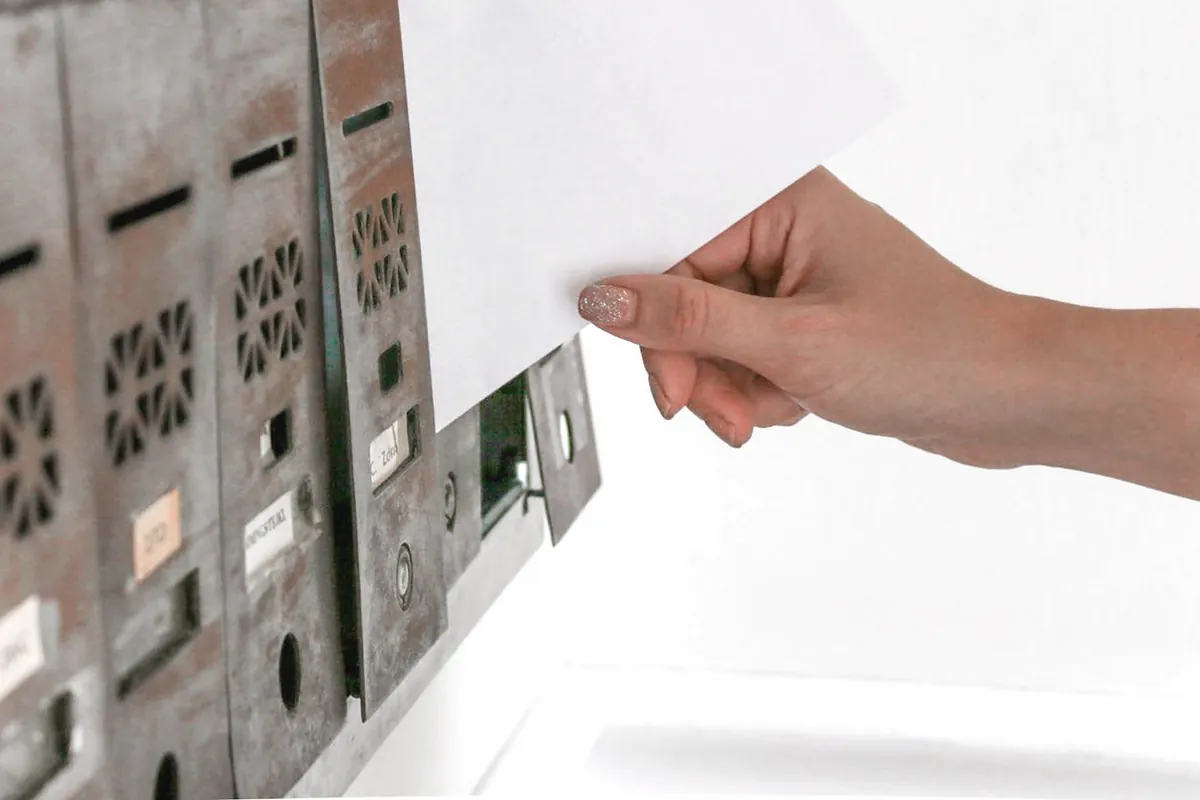 Eine Hand schiebt einen weißen Umschlag in ein Metallfach einer Briefkastenanlage