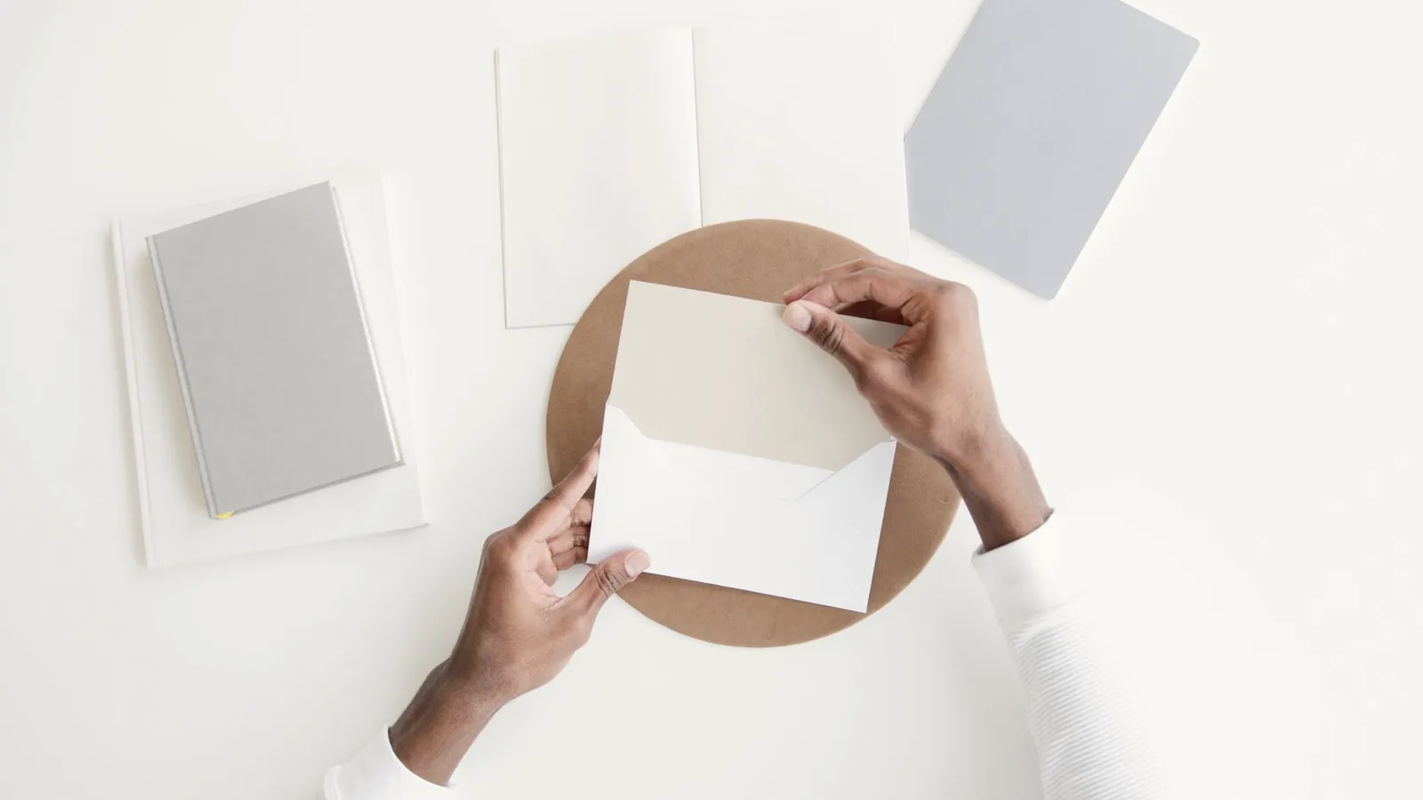 Hände schieben ein weißes Blatt in einen Umschlag, daneben Papier in Pastellfarben