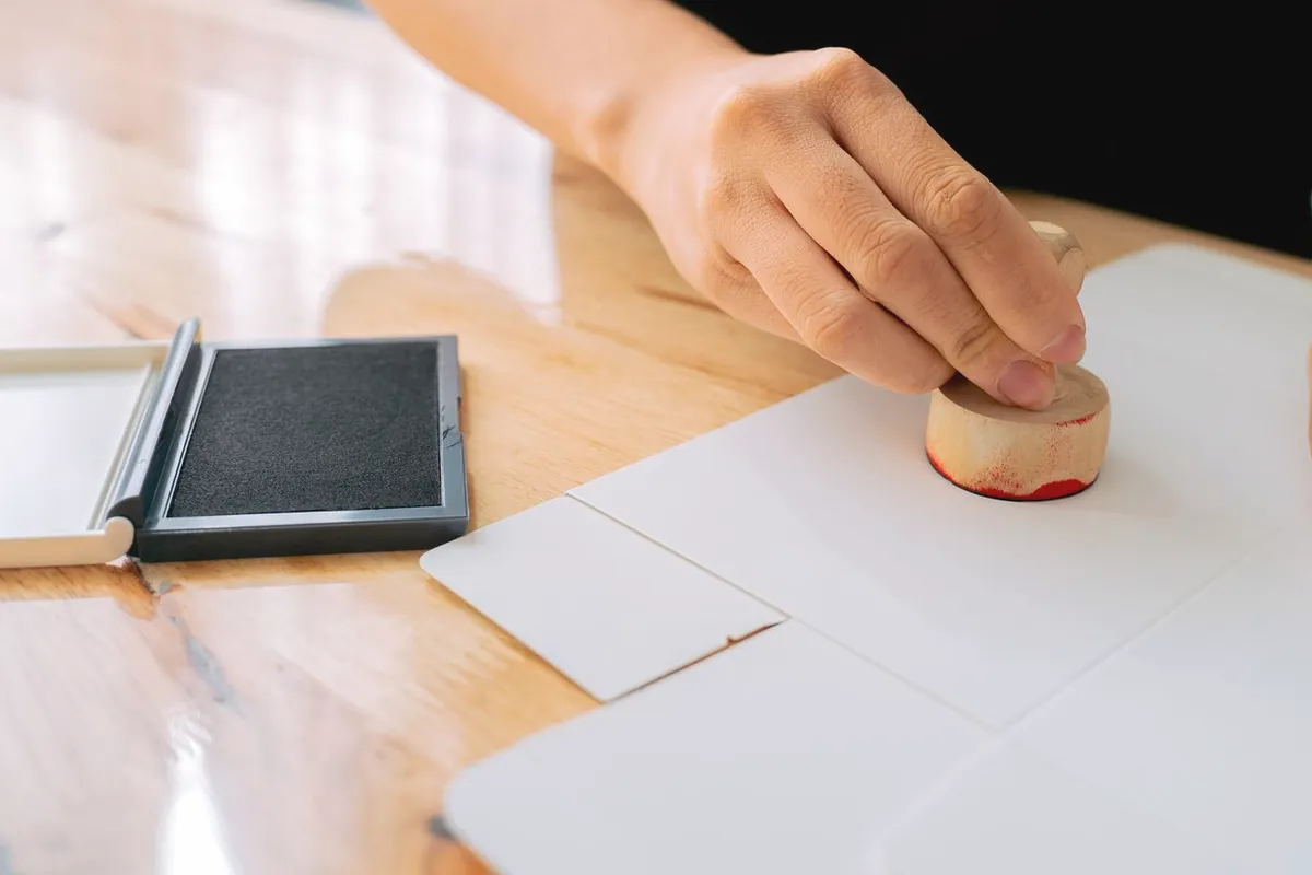 Eine Hand stempelt ein weißes Blatt, daneben ein offenes Stempelkissen